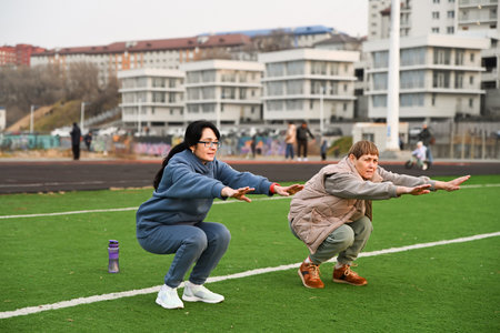 Two women, aged 50 and 60, in tracksuits, doing squats at the stadiumの写真素材