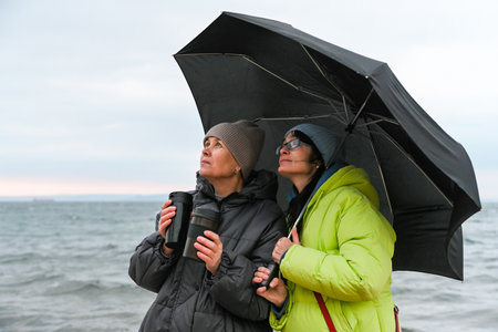 Two Russian women, aged 50 and 60, wearing hats and jackets, hold coffee in a thermos while walking under an umbrella.の写真素材