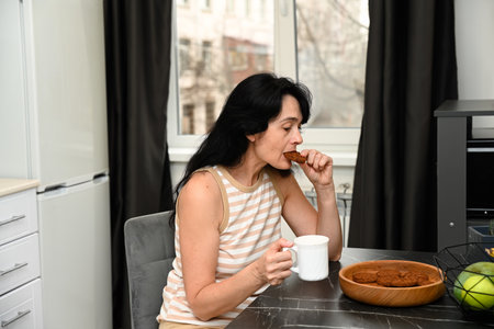 Mature 50 years old Russian woman enjoys a calm sleepy morning holding coffee and a cookie, taking a bite while sitting quietly with a soft peaceful mood.の写真素材