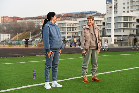 Two women, aged 50 and 60, in tracksuits, doing squats at the stadium and laughingの写真素材