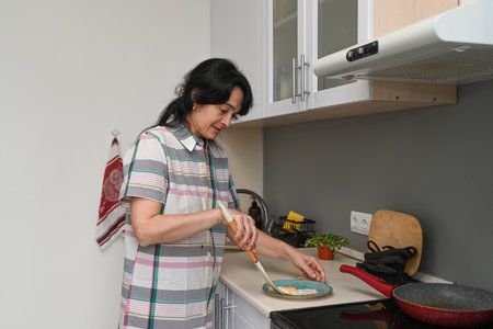 Mature 50 years old Russian woman placing freshly cooked fried eggs on a plate in a cozy home kitchen, showing everyday cooking and simple routine.の写真素材