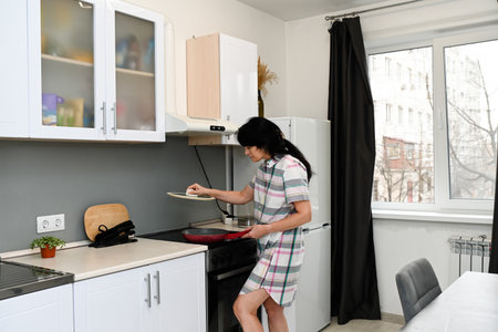 Mature 50 years old Russian woman lifting a pan lid in a bright kitchen, preparing food, looking calm and focused with visible wrinkles and morning moodの写真素材