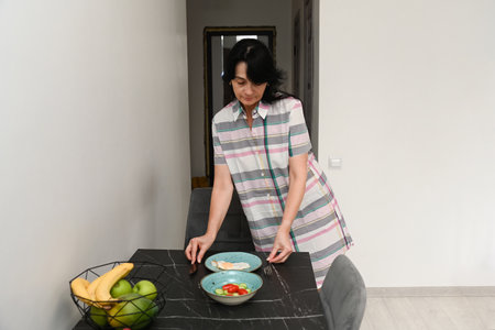 Mature 50 years old woman setting a table with sunny side up eggs and fresh vegetables, showing a simple home breakfast and calm everyday routine.の写真素材