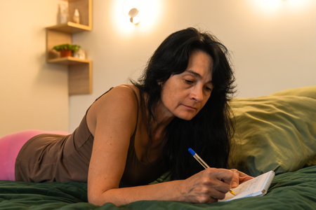 A 50 year old woman lies on her bed writing plans for tomorrow in a notebook, natural look with no makeup, visible wrinkles, evening planning routine.の写真素材