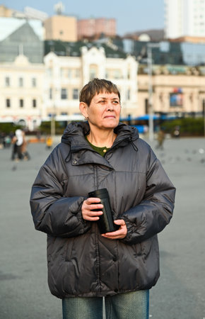 60 year old russian woman with short hair, holding thermos cup with coffee standing in city Vladivostok and looking rightの写真素材
