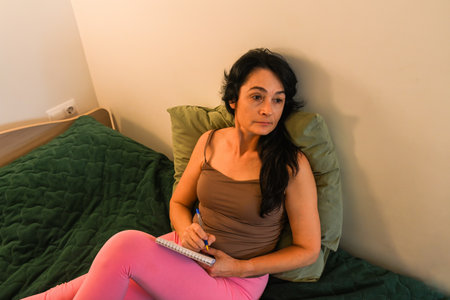 50 year old woman sitting on her bed pausing with her notebook reflecting on her thoughts seeking calm awareness clarity and emotional reliefの写真素材