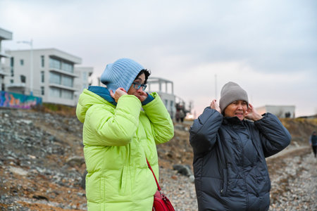 Two Russian women 50 years old and 60 years old adjusting warm hats during a cold coastal walk in Vladivostok showing comfort care and simple everyday connectionの写真素材