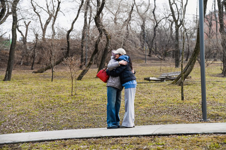 Two women 50 years old and 60 years old share a warm hug outdoors in early spring, expressing support, connection, calm and emotional openness in a quiet park.の写真素材