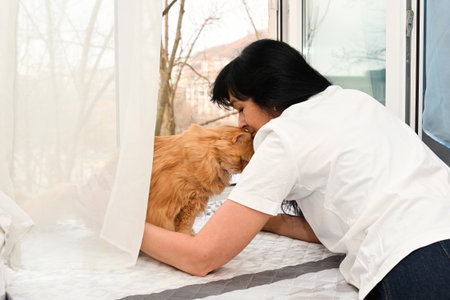 Woman 50 shares a quiet mindful moment with her senior cat by the window creating a sense of warmth trust and emotional comfort in a calm home spaceの写真素材