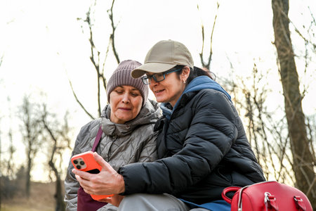 Two women aged 50 and 60 sit together in a quiet park at dusk, calmly sharing a moment while looking at a phone and enjoying a sense of connection and peace.の写真素材