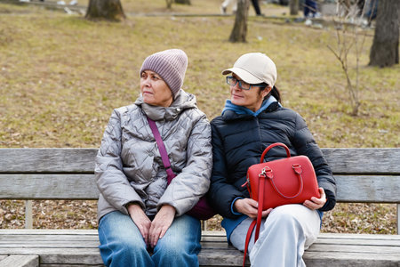 Two friends 50 and 60 years old sit in a spring park looking left enjoying a quiet moment together with calm presence freedom and relaxed connection.の写真素材
