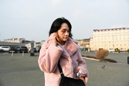 Russian woman 50 years old stands in Vladivostok at sunset, turning on wireless earbuds, showing calm focus, evening freedom, and natural facial wrinkles.の写真素材