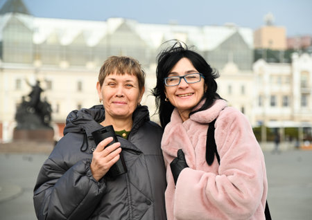 Two Russian friends, 50 and 60 years old, smiling warmly at the camera in the city, sharing joy, confidence, lightness, and the ease of mature female friendship.の写真素材