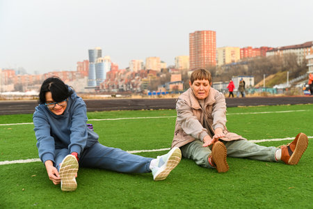 Two Russian women aged 50 and 60 stretch forward while sitting on grass at a stadium in Vladivostok, staying active together and supporting healthy lifeの写真素材