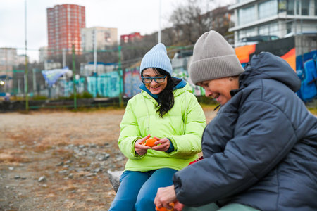 Two women 50 and 60 years old sit outdoors in Vladivostok eating fruit and enjoying calm mindful time together in cool spring weatherの写真素材