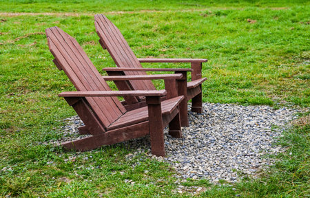 Two wooden Adirondack chairs on a small gravel area in a green grassy field, creating a peaceful outdoor seating background with copy space and nobody around.の写真素材