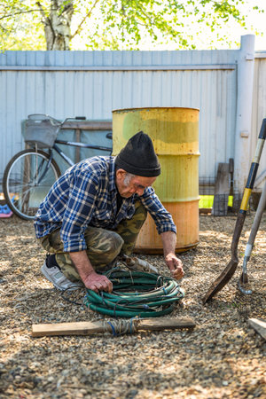 Elderly Russian man crouching near a barrel and checking a hose, surrounded by village tools and a...