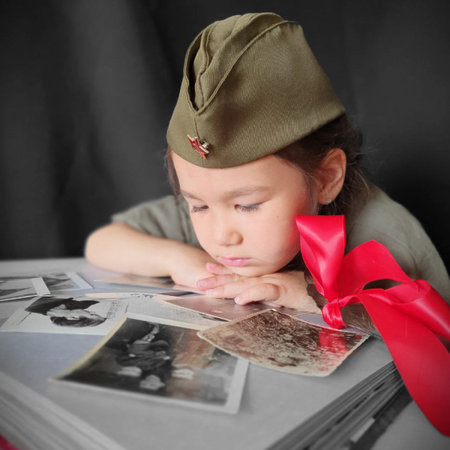 A little girl in a military uniform with two pigtails and red bows is looking at an album with...