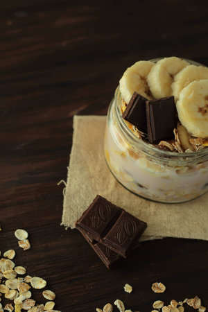 healthy delicious dessert - muesli and yogurt with banana, nuts and chocolate on dark wooden...