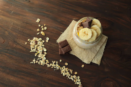 healthy delicious dessert - muesli and yogurt with banana, nuts and chocolate on dark wooden background top viewの写真素材