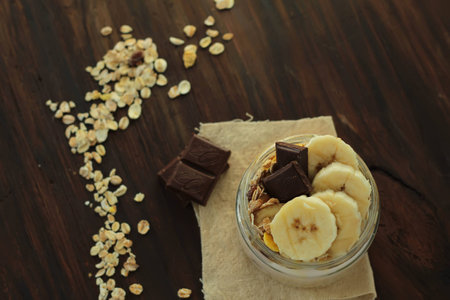 healthy delicious dessert - muesli and yogurt with banana, nuts and chocolate on dark wooden backgroundの写真素材