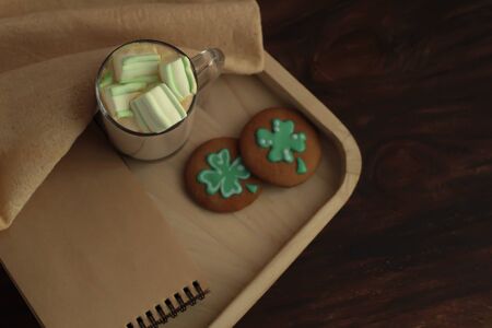 Homemade gingerbread Gourmet St Patrick Day cookies, notebook and a mug of coffee with marshmallows on dark wooden background St Patrick day celebration place for textの写真素材