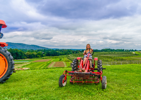 Beautiful woman on a red agricultural tractor, green field, landscape of meadow with grass Upstate New Yorkの写真素材