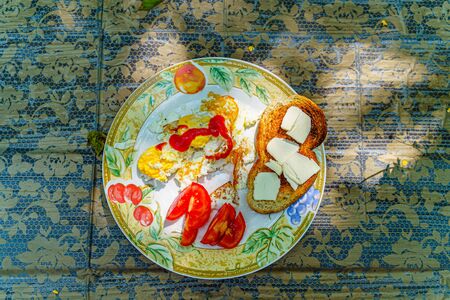 Full breakfast - fried eggs, ketchup, sliced tomato, toasted slice of white bread with slices of butter on a white plate with an ornament on a wooden table covered with a tablecloth, top view.の写真素材