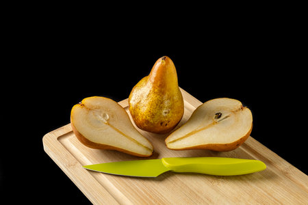 One whole, two halves of a pear and a colored knife on wooden cutting board isolated on black backgroundの写真素材
