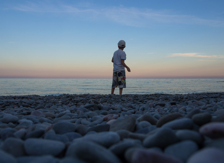 Boy stands in the evening on the beach and throws stones into the waterの写真素材