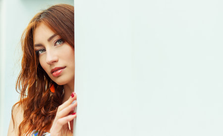 sexy beautiful girl with red hair and full lips peeping from behind the white wall