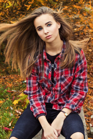 beautiful charming young attractive girl with large blue eyes, with long dark hair in the autumn forest sits on a tree in black pants and shirtの写真素材