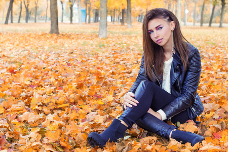 beautiful girl in rock style with bright makeup in white jacket and black pants and boots in a leather jacket sitting in the Park on the yellow leaves in the autumn warm dayの写真素材