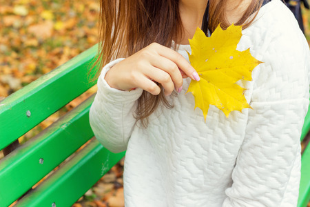 beautiful girl in a white jacket and black pants with yellow leaves in the hands of sitting on the bench in autumn Parkの写真素材
