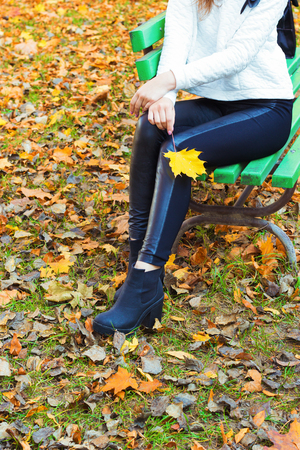 beautiful legs girl in black pants and boots with yellow leaves in the hands of sitting on the bench in autumn Parkの写真素材