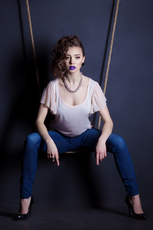 beautiful young sexy girl on a swing from ropes in the Studio on a black background in jeans and bright makeupの写真素材