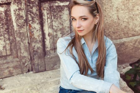attractive beautiful gentle girl sits in the city on the steps of the old building in jeans and fashion footwearの写真素材