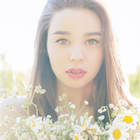 beautiful sexy cute girl with big lips and red lipstick with bared shoulders with a bouquet of daisies at sunset on a sunny warm summer day, the rays of light into the cameraの写真素材