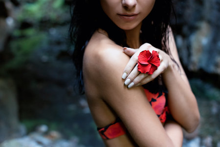 beautiful sexy cute girl with black hair in a wet bathing suit with a flower in her hair and a ring on his finger in the dark rainforest among the rocksの写真素材