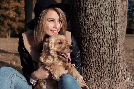 Beautiful cute happy girl in a black hat playing with her dog in a park in autumn another sunny dayの写真素材