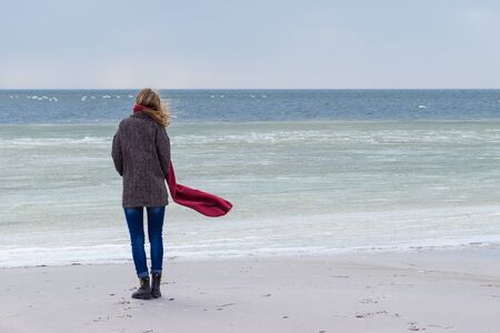 Lone sad beautiful girl walking along the shore of the frozen sea on a cold day, rubella, chicken with a red scarf on the neckの写真素材