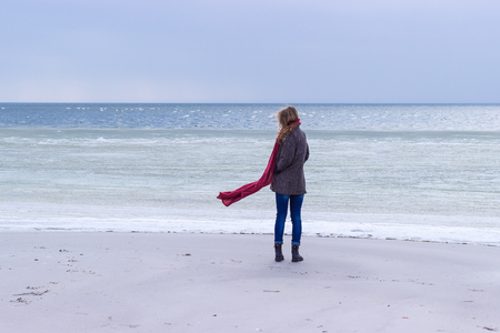 Lone sad beautiful girl walking along the shore of the frozen sea on a cold day, rubella, chicken with a red scarf on the neckの写真素材