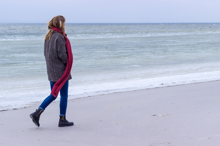 Lone sad beautiful girl walking along the shore of the frozen sea on a cold day, rubella, chicken with a red scarf on the neckの写真素材