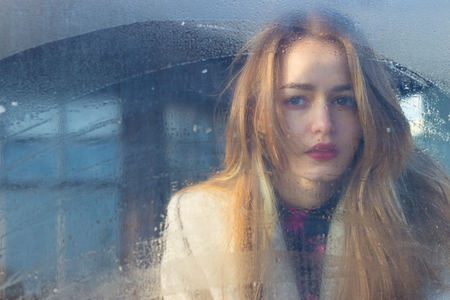 sad beautiful seksalnaya Pretty sad lonely girl behind wet glass with big sad eyes in a coatの写真素材
