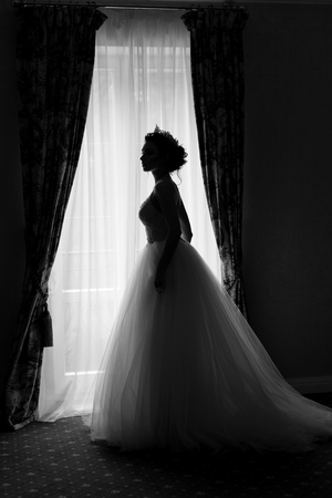 beautiful delicate sexy bride happy woman with a crown on her head by the window with a large wedding bouquet in a luxurious white dressの写真素材