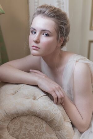 gentle portrait of a young beautiful girl in a light chiffon dress with light make-up and hairstyle sitting on the couch photos in gentle tones in the style of fine artの写真素材
