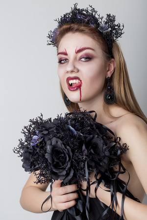 Beautiful cheeky girl in the image of a vampire with bright dark makeup, black vampire bride with a bouquet and a black wreathの写真素材
