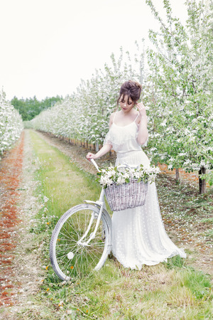 beautiful gentle lovely girl bride in a white dress standing with his bicycle in blossoming apple orchardの写真素材
