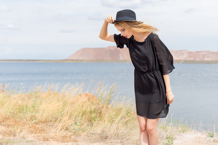 beautiful young blond woman in a black dress and a light black hat in the desert and the wind blowing her hair in a hot summer dayの写真素材