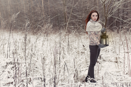 beautiful red-haired girl in mittens walking with a lamp in a fabulous winter forestの写真素材